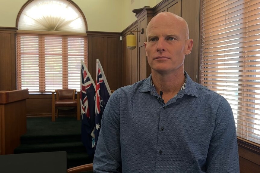 A man in a blue shirt stands in a council chambers in front of two Australian flags 