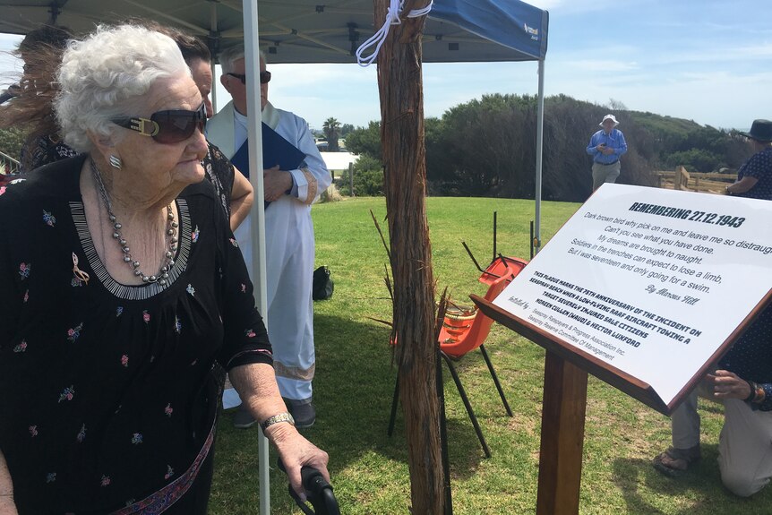 An elderly woman leans on a cane and looks at a plaque. 