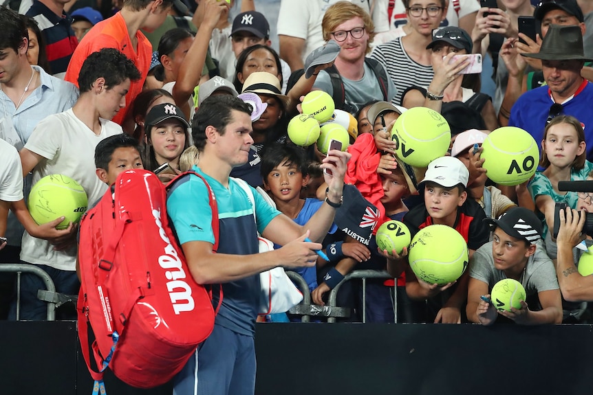 A male tennis player holds a mobile phone to take a picture, as fans behind him hold giant tennis balls for autographs.