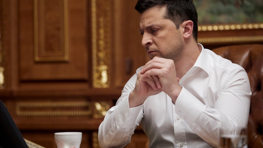 Volodymyr Zelenskyy sitting at his desk in a white shirt listening intently.