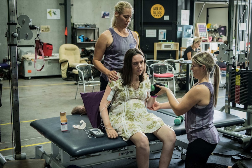 A woman kneels behind Jess Collins, while another assists her lift a small weight in a gym.