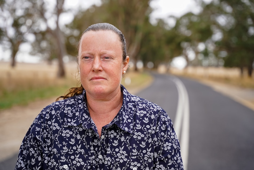 A women wearing a blue buttoned shirt stands in the middle of a country road looking out, reflective.