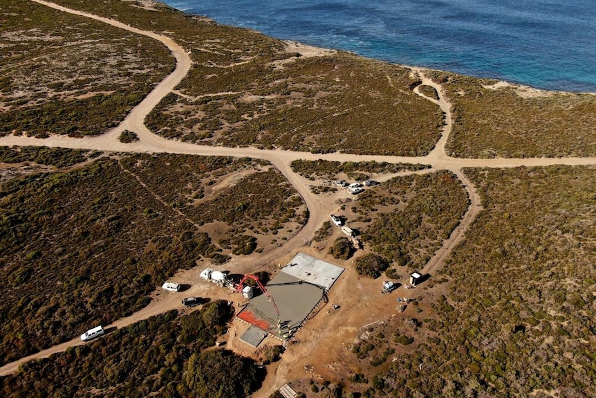 Drone vision of a concrete pad with large crane-like machinery at one end. Pad is surrounded by grasslands and near the beach 