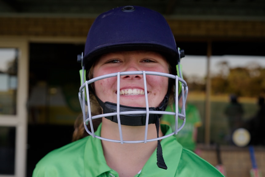 A young woman with fair skin smiles at the camera, wearing a cricket helmet 