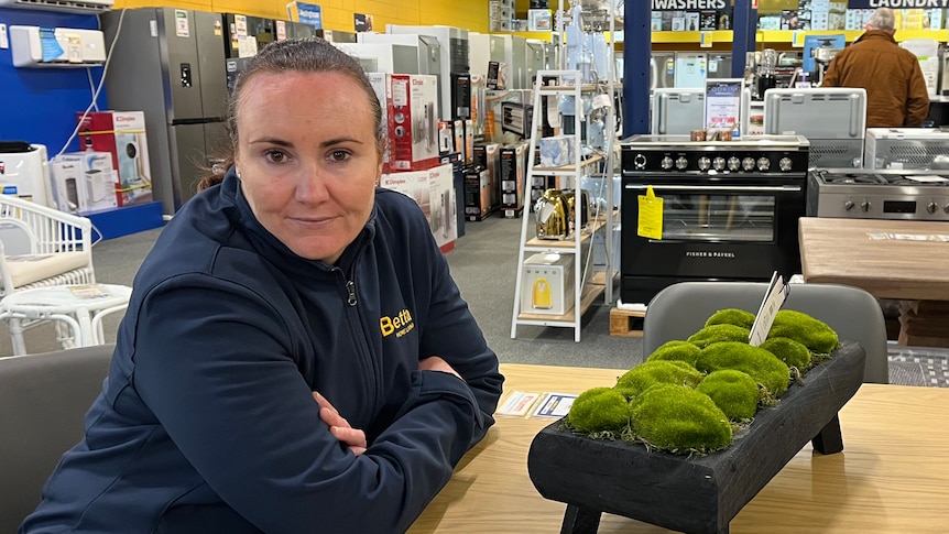 A lady in a blue puffer jacket sits as a wooden table with her arms crossed in an electrical store
