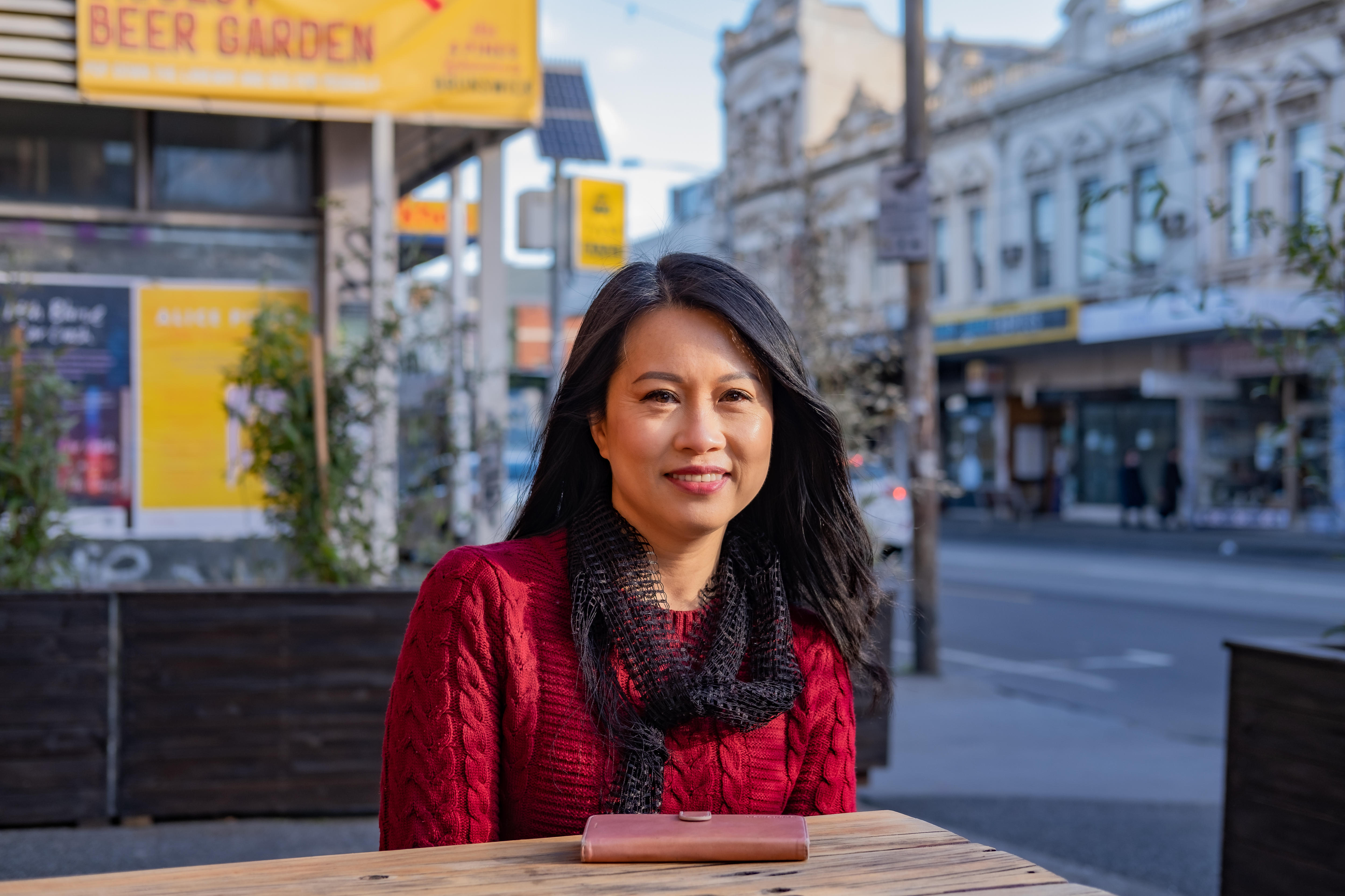 A woman sits at a table and looks into the camera smiling.