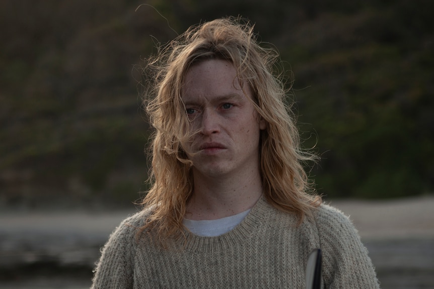 Young man with wavy blonde hair stands on a beach wearing a taupe knitted jumper, holding a surfboard and looking disgruntled.