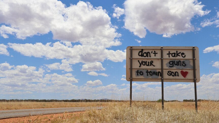 Blue sky, fluffy clouds, above a highway and red dirt. Sprayed on a road sign is "don't take your gun to town son"