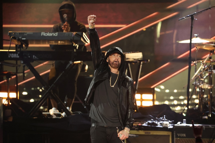 Eminem stands on stage dressed in all black with his arm raised in the air.