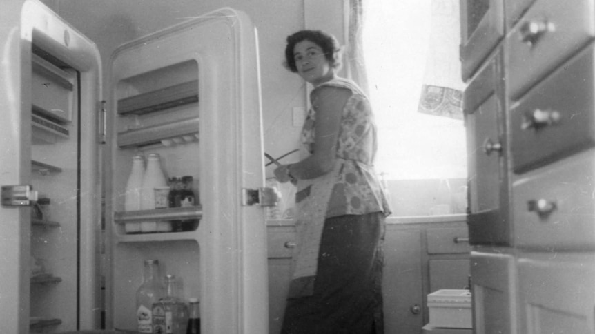 Woman in 1950s kitchen with fridge with open door.