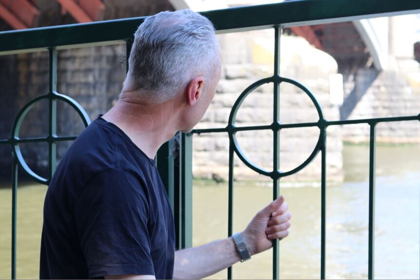 A man crouches against a green railing in black t-shirt, looking out over water.