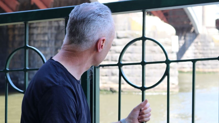 A man crouches against a green railing in black t-shirt, looking out over water.