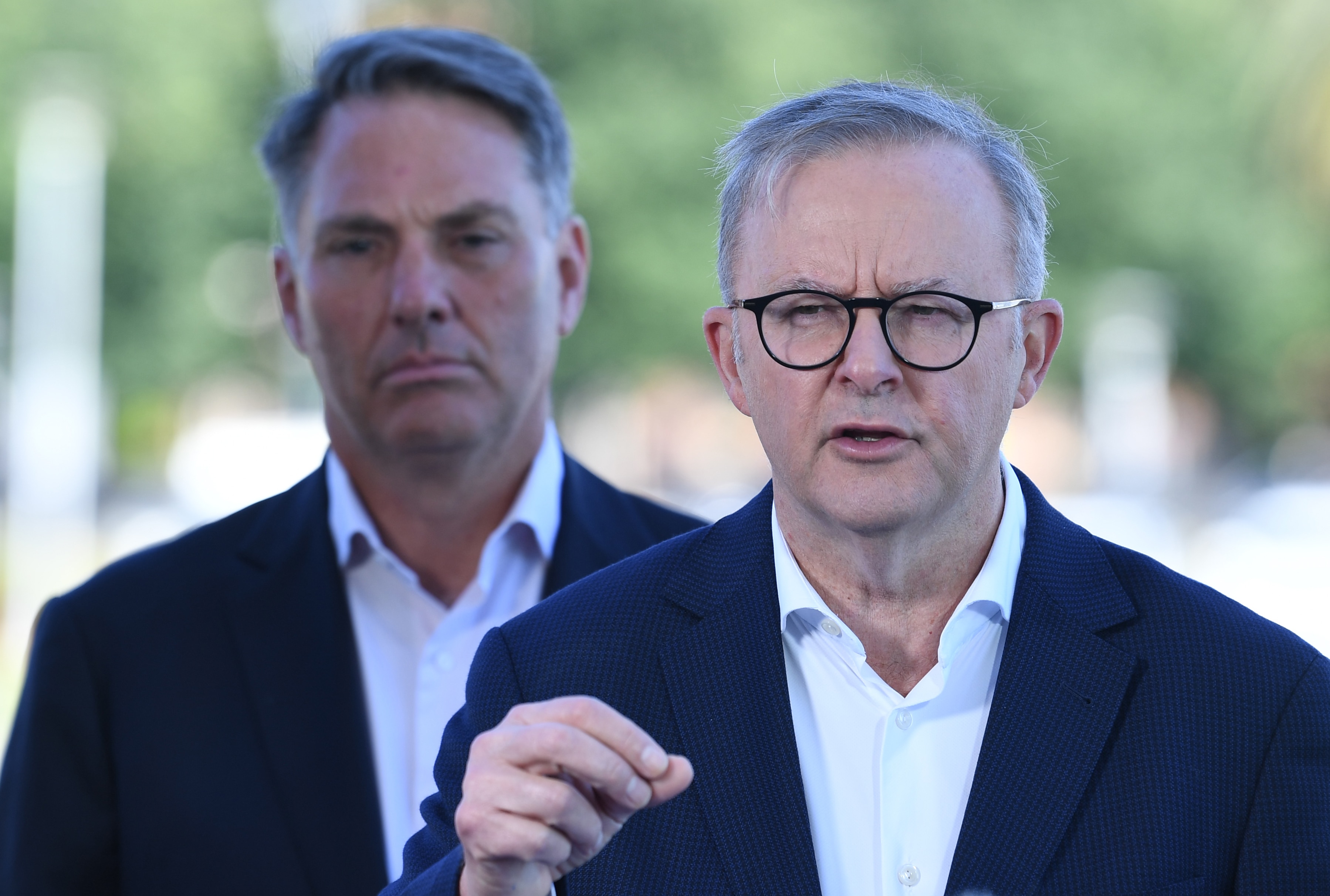Mr Albanese stands wearing a suit and is speaking with Mr Marles in the background. 