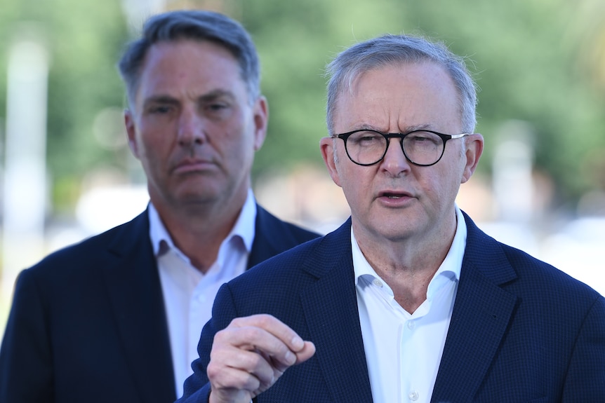 Mr Albanese stands wearing a suit and is speaking with Mr Marles in the background. 