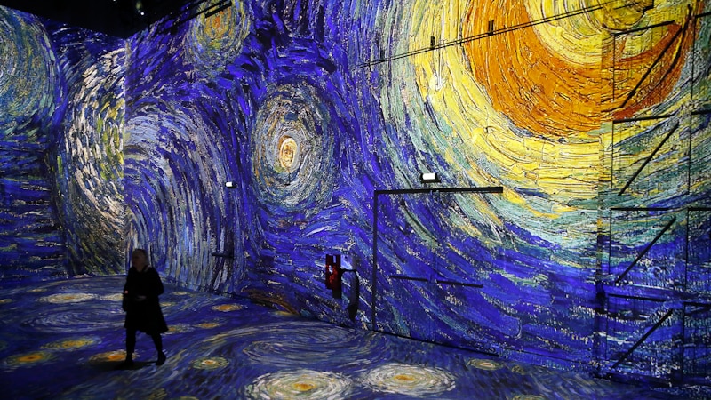 Darkened gallery space with projections of Van Gogh's starry night on floor and walls, and figure walking through space.