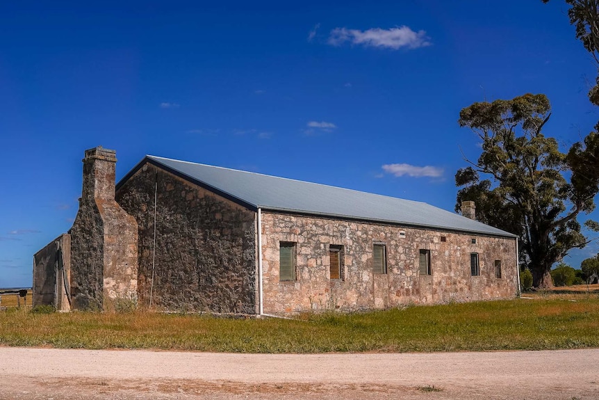 A long old stone cottage under a bright blue sky next to gum trees.