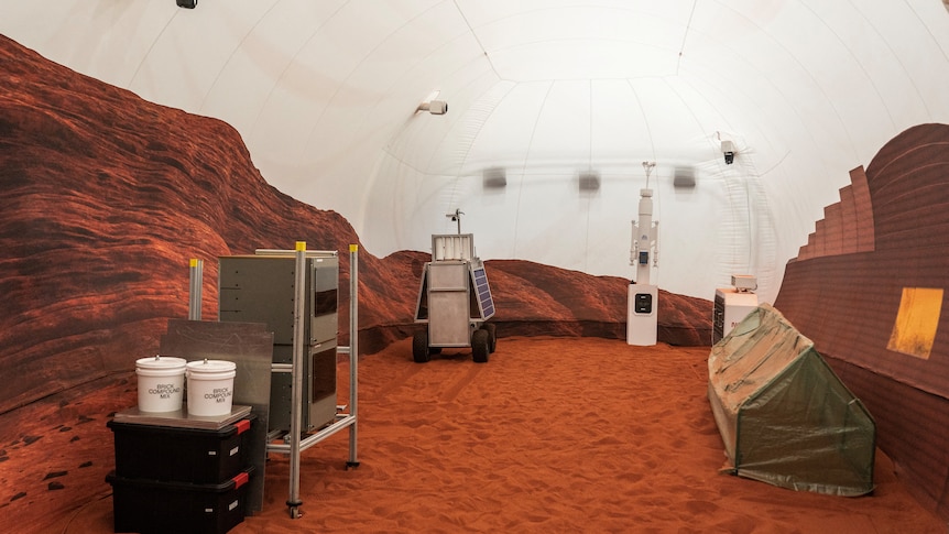 A room with red sand on the floor, on the left and right side is machinery. On the walls are printed red rocks.