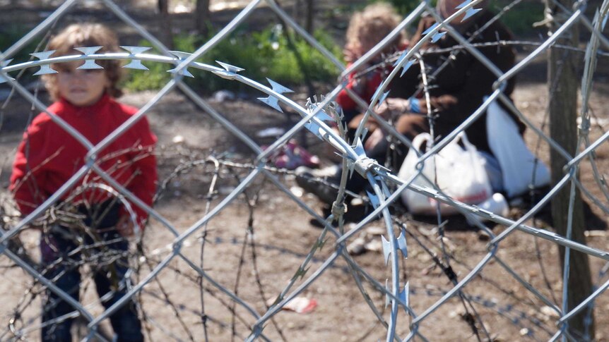A Syrian child, out of focus, stands next to razor wire.