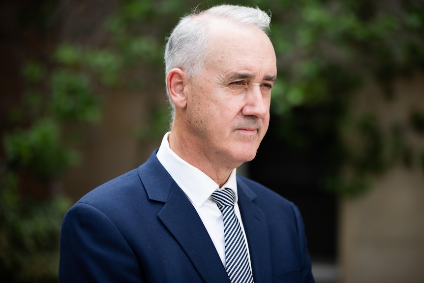 Man in blue suit and white shirt stands with a contemplative expression.