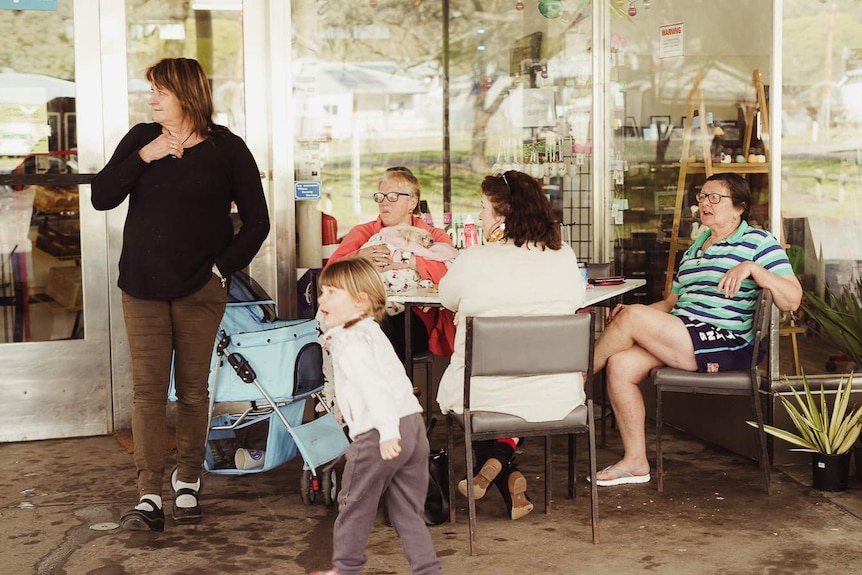 A woman stands next to a table of women chatting sitting down outside a glass shopfront.