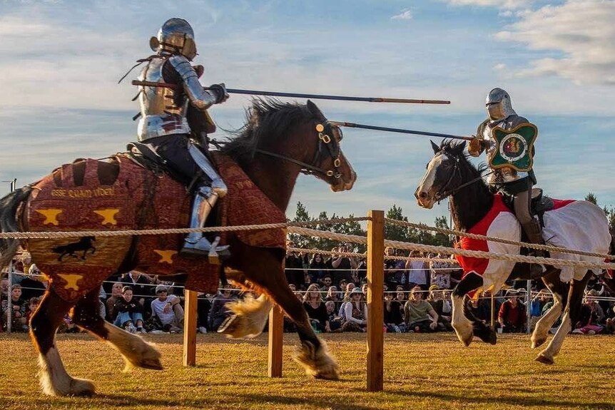 Two people dressed as knights hold long lances while facing each other on horseback.
