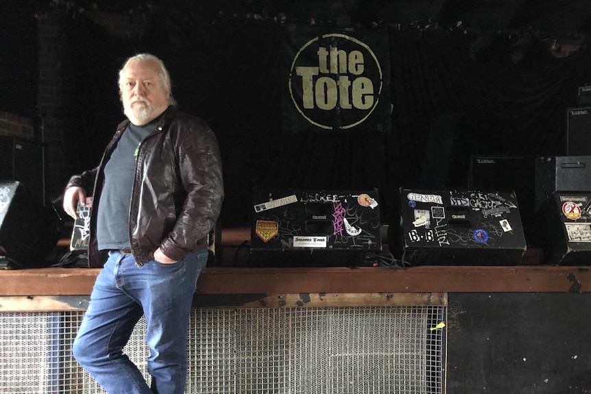 A beareded man standing in a dark room next to a sign The Tote.