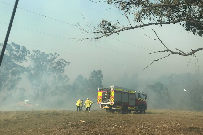 Firefighters in yellow jackets stand next to a red fire truck on grass surrounded by smoke.