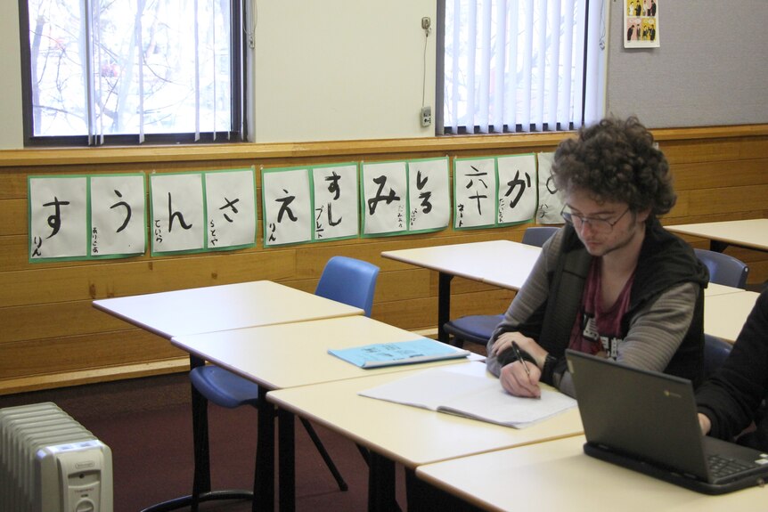 A male student sits at a desk in a mostly empty classroom. Asian calligraphy lines the wall behind him.