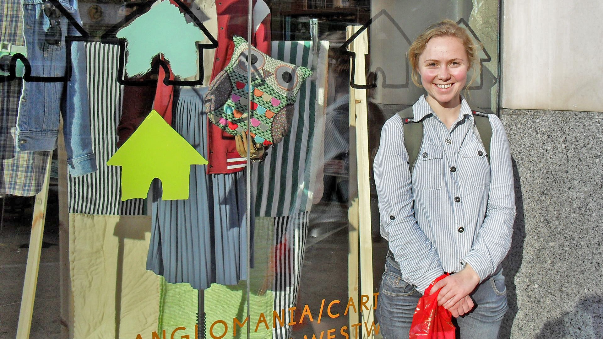 A smiling Daisy stands in front of a Paris shop window displaying clothes and an owl handbag