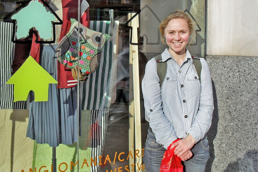 A smiling Daisy stands in front of a Paris shop window displaying clothes and an owl handbag