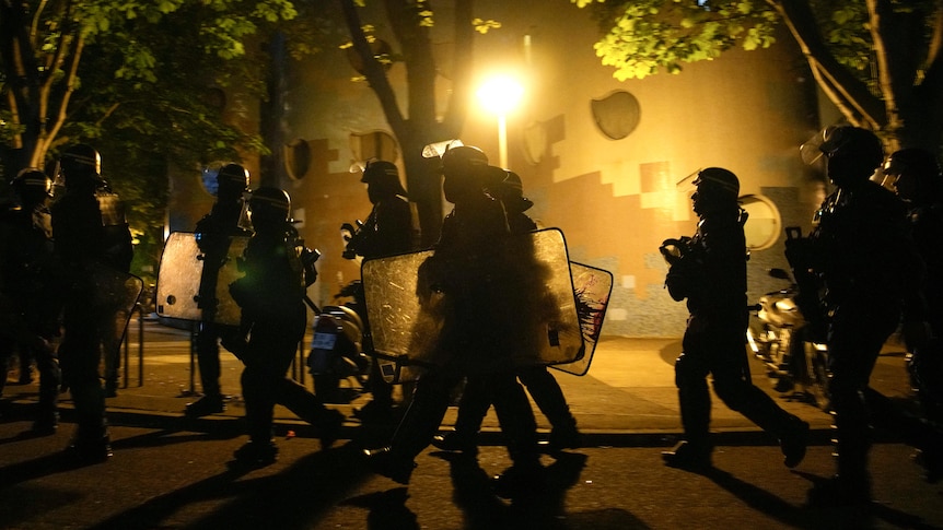 A group of helmeted police officers walk with riot shields during a protest, lit by bright yellow street light.