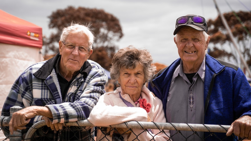 Three smiling elderly people leaning against a fence, looking at the camera. Two men and a woman who is in the middle.