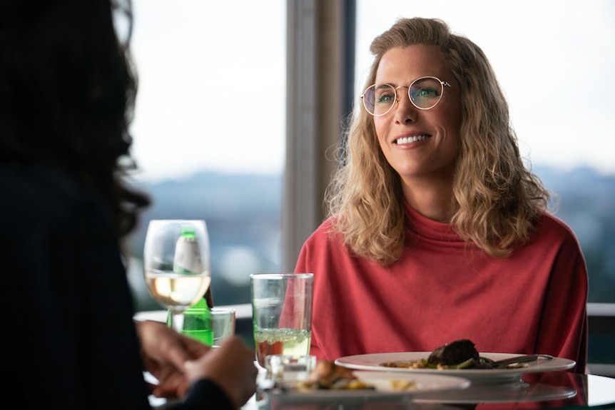 Kristen Wiig wearing red jumper and gold-rimmed round glasses, smiling, sitting at table across from someone with meal and drink