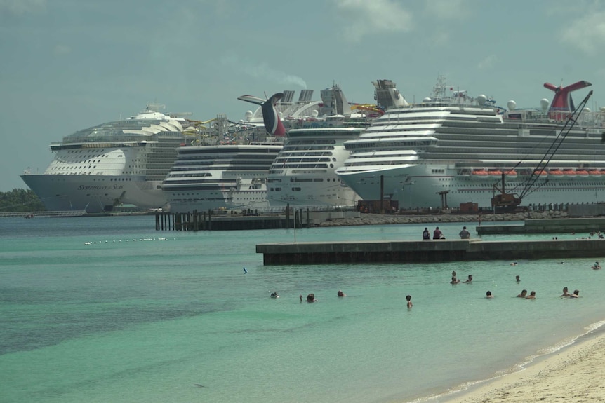 In the distance, large cruise ships are docked. In the foreground people swim in the ocean.