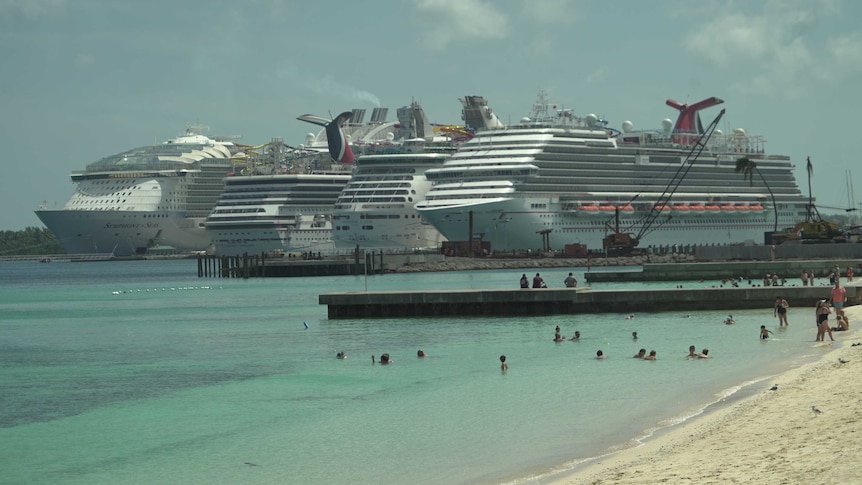 In the distance, large cruise ships are docked. In the foreground people swim in the ocean.