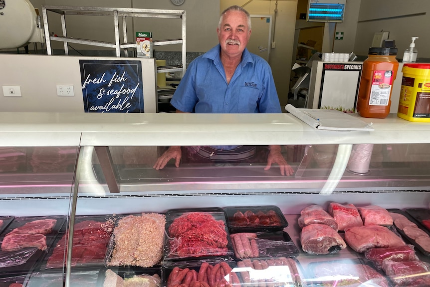 A white & grey haired middle aged man grins in a butchers apron in front of a display case of different cuts of meat 