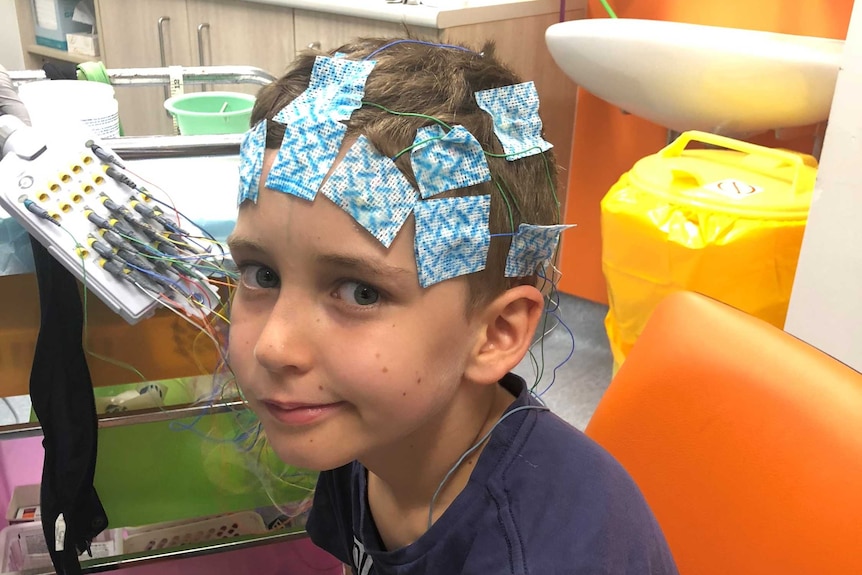 Lewis Railton smiling to camera with patches and wires on his head at the hospital.