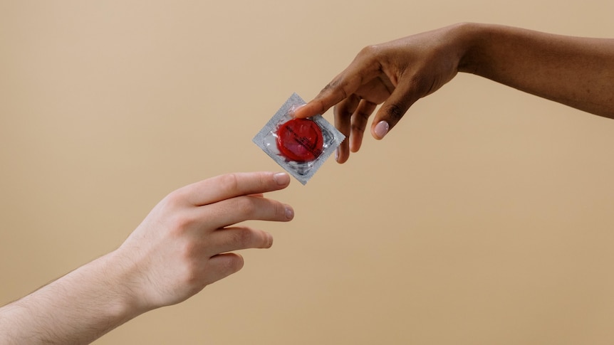 Two hands reach out and pass a condom between them against a beige plain background.