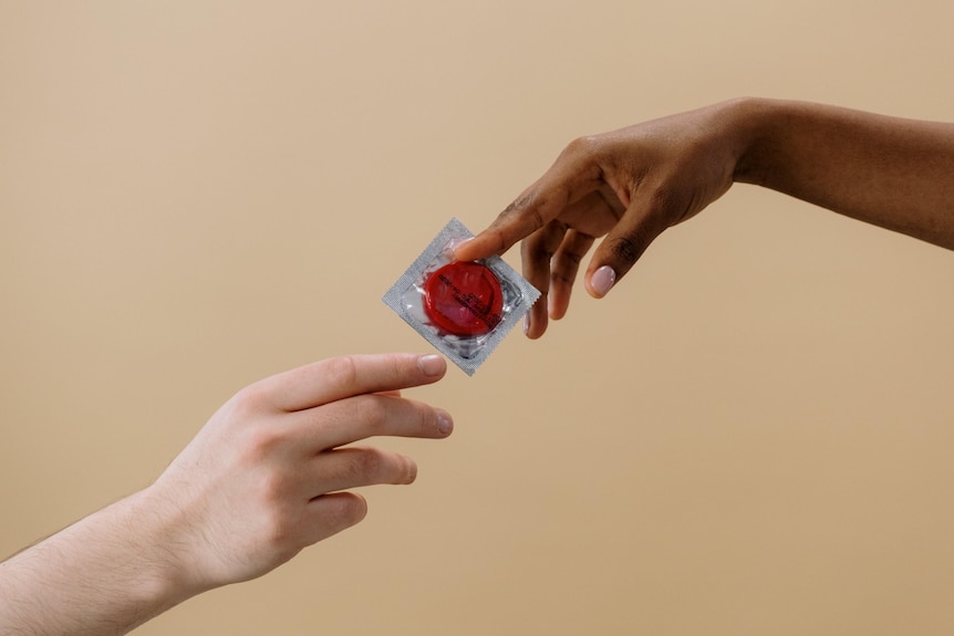 Two hands reach out and pass a condom between them against a beige plain background.