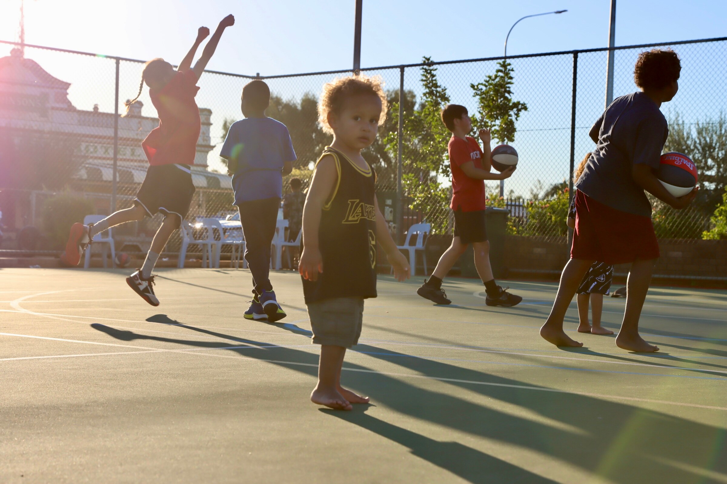 A young Aboriginal boy looking at the camera with older children playing basketball in background.