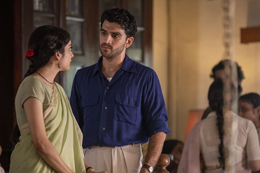 A man with black hair and a beard wearing a dark blue shirt stares at a woman with black hair and wearing a green sari