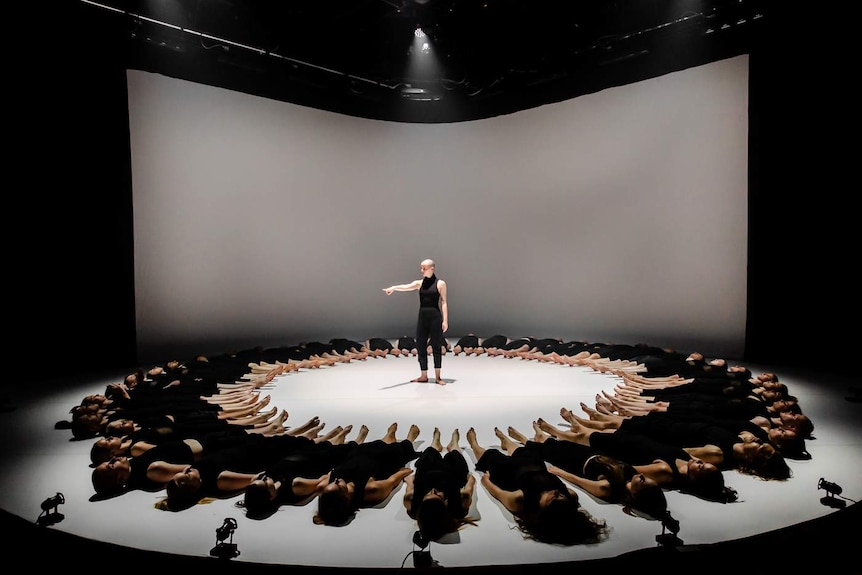 Circular stage on which circle of 49 dancers dressed in black lie with feet pointed to centre, and solo dancer in middle.