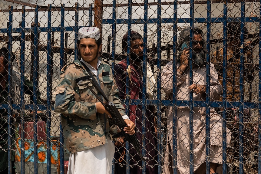 A man with a gun across his chest stands against a metal fence. 