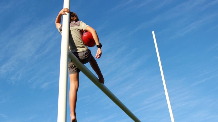 A boy sits balancing on football goalposts with a footy in his hand.
