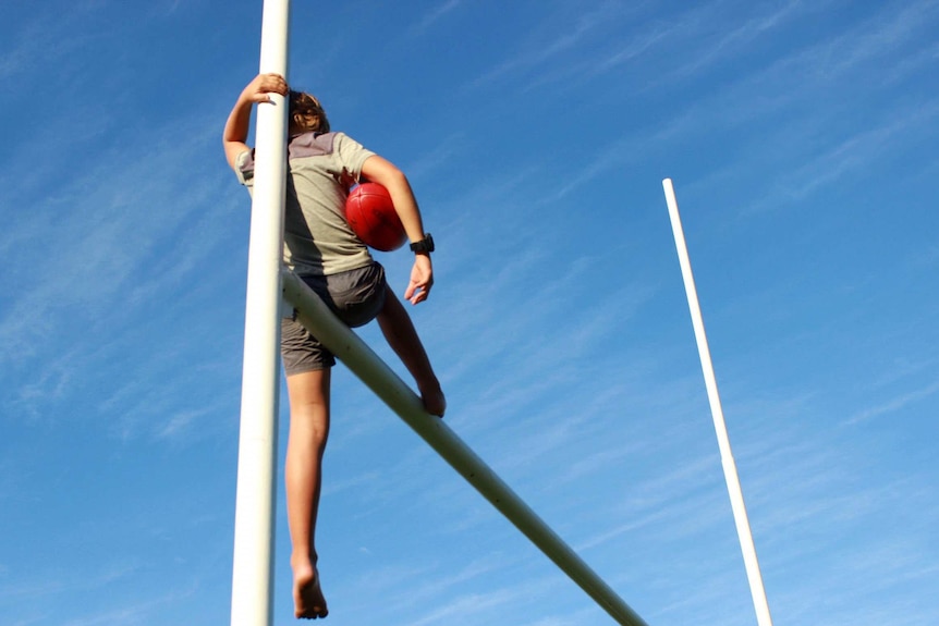 A boy sits balancing on football goalposts with a footy in his hand.