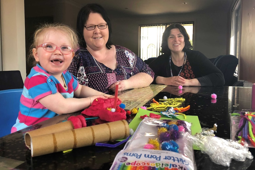 A little girl sits at t table with two adults, smiling. She is playing with plasticine and other craft materials, like pom poms.