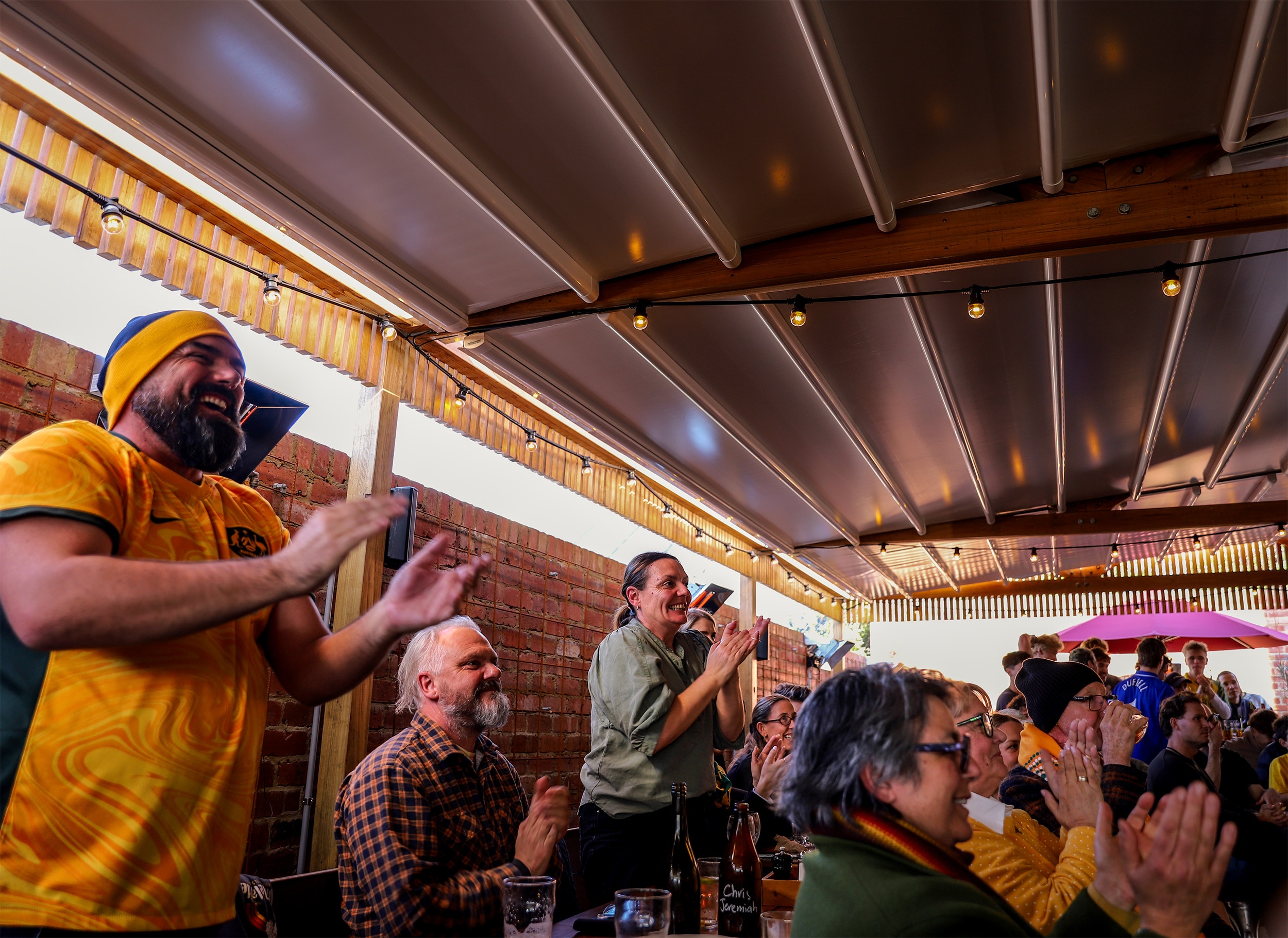 A beer garden of people cheering, some wearing yellow Australian outfits, amid wooden tables and brick wall