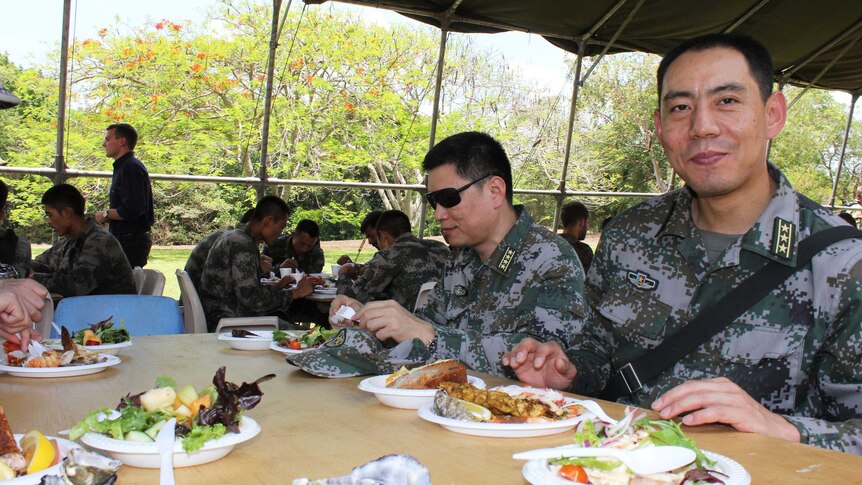 A Chinese military personnel sits with other troops at a table loaded up with plates of food.