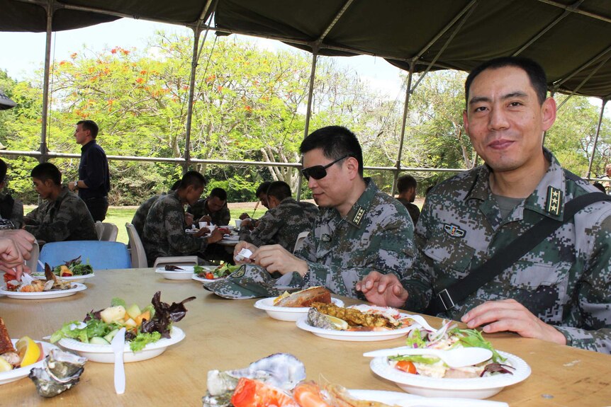 A Chinese military personnel sits with other troops at a table loaded up with plates of food.