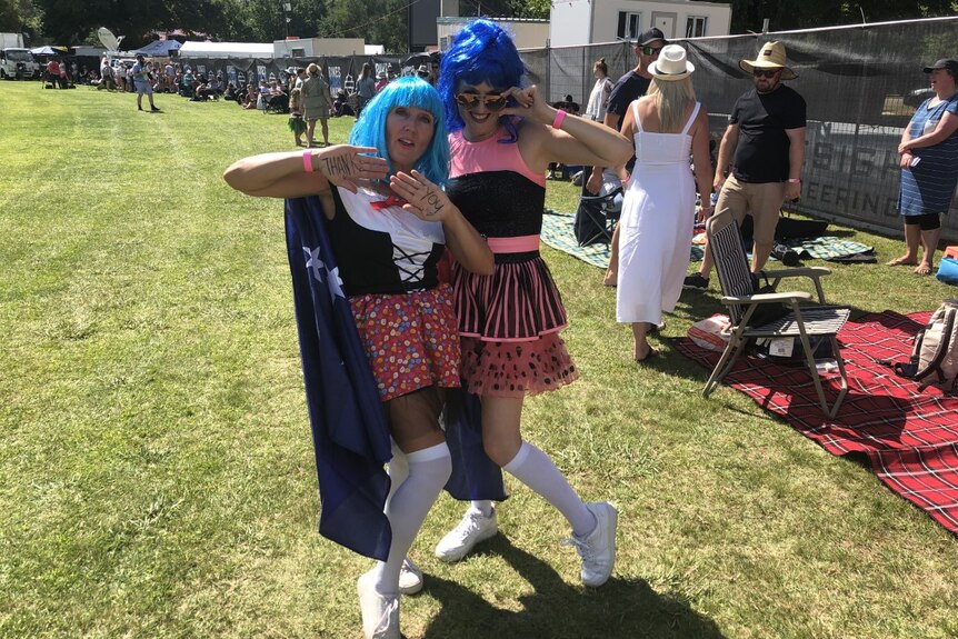 Two women stand on grass posing in brightly coloured outfits and blue wigs with a crowds behind them on a sunny day.